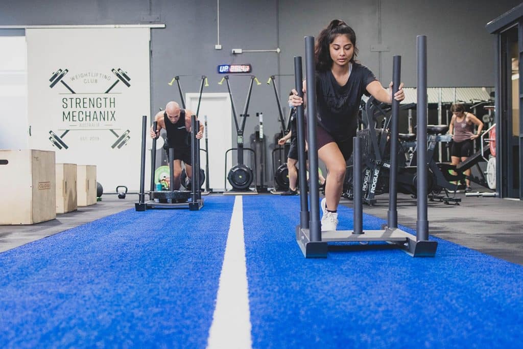 Two people kickstart their fitness by pushing weighted sleds along a blue turf strip in a gym. Gym equipment and a Strength Mechanix sign are visible, while both individuals appear focused on their workout.