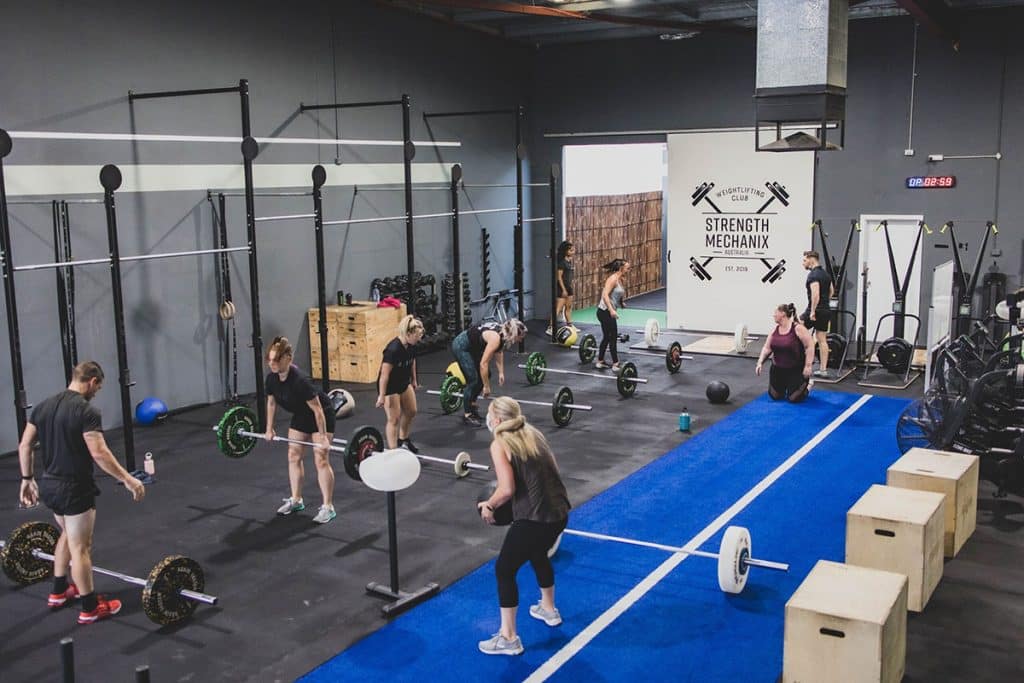 A group of people kickstart your fitness journey with barbells in a gym with high ceilings, wall racks, wooden boxes, and fitness equipment. A sign on the wall reads "Strength Mechanix.