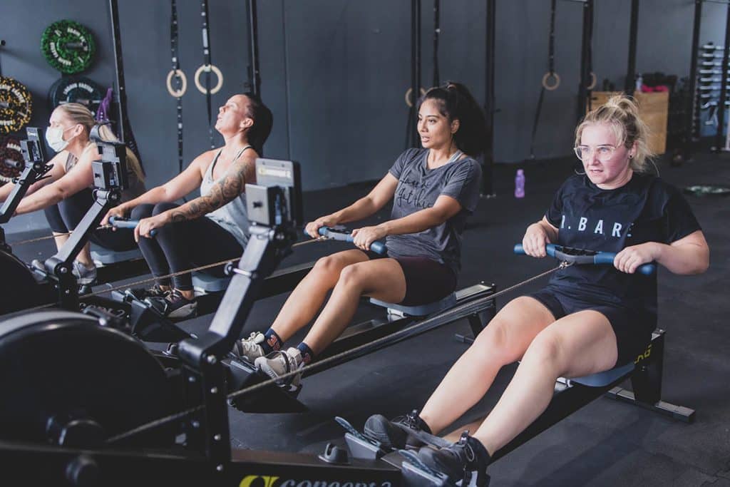 Four women are exercising on rowing machines in a gym, focused and working hard to Kickstart Your Fitness. Gym equipment and fitness rings are visible in the background.